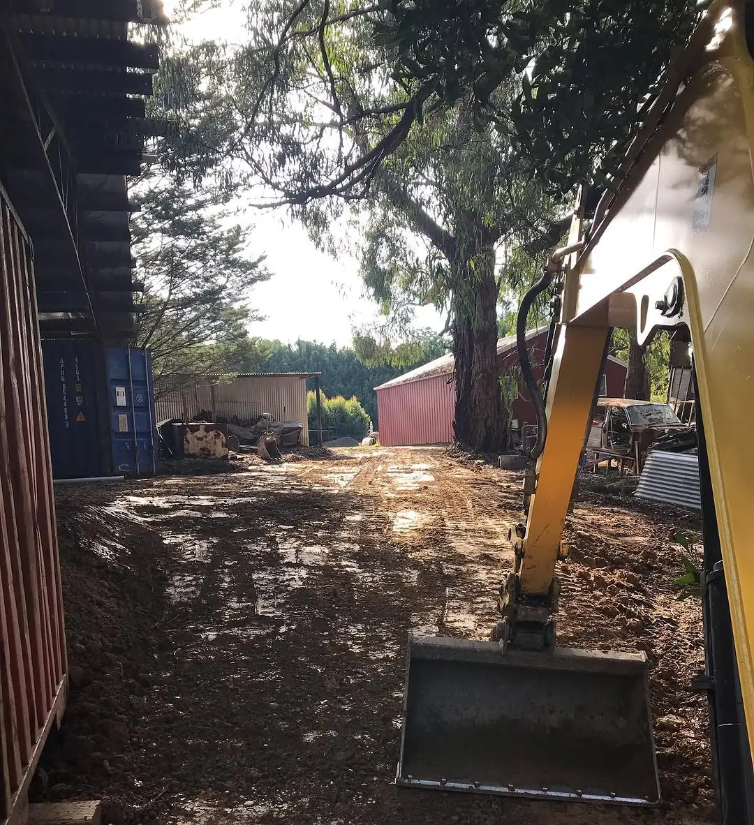 Excavator bucket in the foreground of a freshly cut, wet dirt driveway at Monbulk, running between shipping containers and a red shed